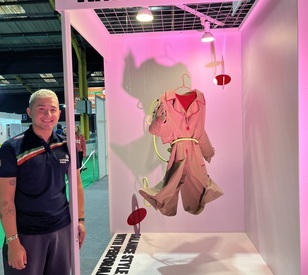 A student stands beside a fashion display featuring a pink and beige outfit suspended against a pink backdrop at an exhibition.