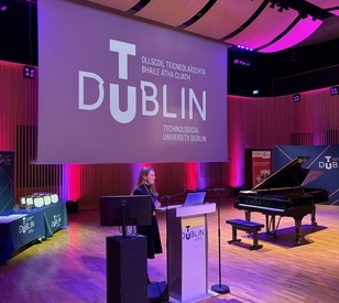 A speaker stands at a lectern on a stage at TU Dublin, with a grand piano and a large screen displaying the university’s branding in the background.
