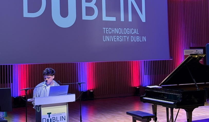 A speaker addresses an audience from a lectern beneath a large TU Dublin-branded screen, with a grand piano on stage to the right.