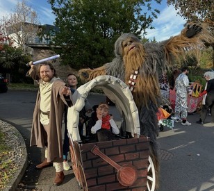 A person dressed as a Jedi, holding a lightsaber, stands alongside a child and a participant dressed as Chewbacca, with a homemade wooden-style cart in the foreground.