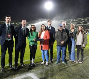 A group of eight adults stand together on a wet football pitch at night, smiling for a photo under bright stadium lights. One woman in the center holds a small trophy or presentation item.