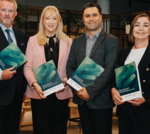 Four professionally dressed adults stand together indoors, each holding a teal-and-green booklet or award, posing for a group photo in a modern office with plants and wood furnishings.