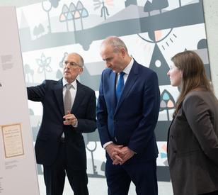 Three people standing at an indoor exhibition, with  one person     gesturing towards an information display while the two others listen.