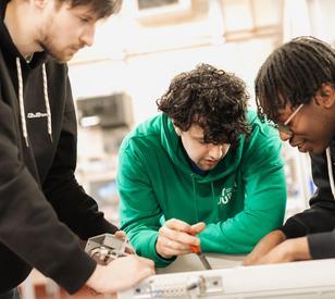 Three students collaborate on a hands-on electronics project, leaning over a workbench and examining a circuit board and equipment together.