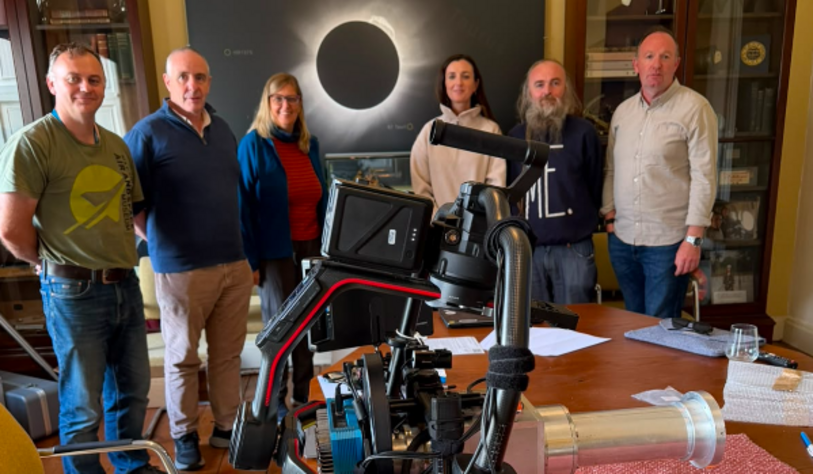 Six people stand behind a camera setup, with a large image of a total solar eclipse displayed on a screen in the background.