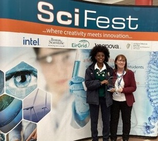 Two people stand smiling with an award in front of a SciFest backdrop. Keywords include: creativity, innovation, and science. The mood is celebratory.