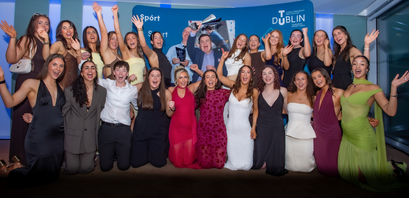 A group of smiling people pose together, raising their arms in celebration. They are dressed formally, conveying joy and excitement. A backdrop displays