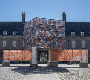 A large building with a cube-shaped structure covered in patterned fabric and orange mesh panels, set in a cobblestone courtyard under a clear blue sky.