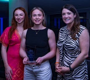 Three women are standing indoors, smiling. The center woman, in a black top, holds a glass award. The others wear a pink dress and a zebra-patterned dress.
