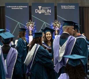 Graduates in teal caps and gowns celebrate at a ceremony. One woman, smiling, stands out amidst the group. Background banners read 