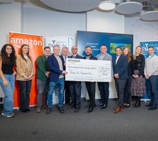 A group of people smiling and standing in a row indoors, holding a large check for thirty-five thousand euros from Amazon to Technological University Dublin. Various banners are visible in the background.