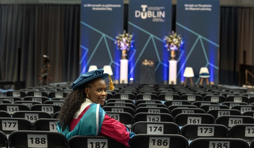A graduate in a cap and gown smiles while seated among numbered chairs facing a stage with floral arrangements. The backdrop reads