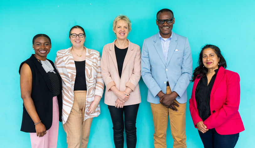 Five diverse people stand smiling against a bright turquoise background. They're dressed in business-casual attire, conveying a friendly and professional tone.