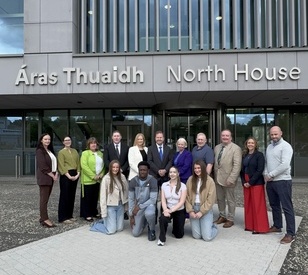 A group of fifteen people stands in front of a building entrance with the sign “Áras Thuaidh North House” above them, posing for a group photo.