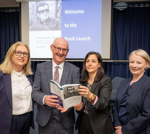 Four people stand smiling, holding a book, in front of a presentation screen at a book launch event. The atmosphere is formal and celebratory.