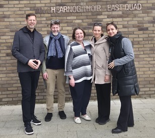 Five people stand closely together, smiling against a brick wall background. Their relaxed poses and warm smiles convey a sense of friendship and camaraderie.
