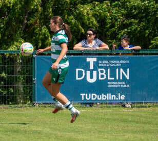 A female soccer player jumps to control a ball mid-air on a sunny day. A blue TU Dublin banner and spectators are in the background. The scene is energetic.