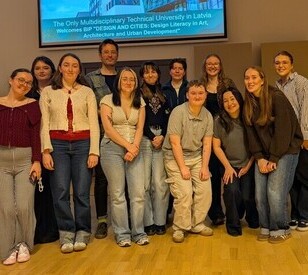 A diverse group of people stands smiling on a wooden floor in front of a screen displaying a welcome message. The atmosphere is warm and cheerful.