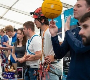 Busy indoor event with people chatting at a booth; a climber in a helmet next to an orange ball, creating a lively, friendly mood.