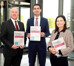 Three people in formal attire stand holding documents, smiling. They are in a modern office with large windows, suggesting a professional setting.