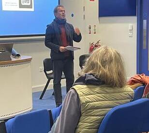 A man in a scarf speaks in front of a seated audience in a classroom. A screen displays a QR code. The atmosphere is engaging and attentive.