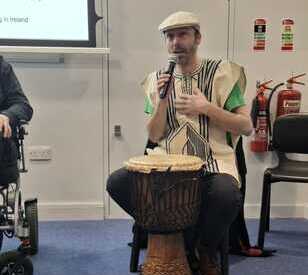 Person seated, playing a djembe drum, wearing a decorative tunic and flat cap, engages audience with a microphone in a casual setting.
