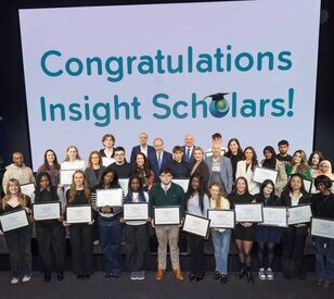 A diverse group of people stands smiling, holding certificates. Behind them, a large screen displays 