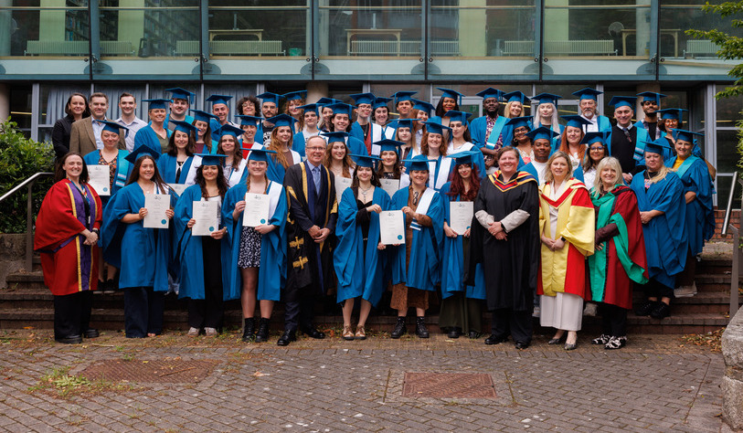 A group of graduates in blue gowns and caps, holding diplomas, stand with faculty in colorful robes. They smile in front of a modern building.