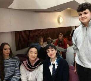 A group of four young adults sitting together, smiling in a cozy indoor setting. The atmosphere is relaxed and friendly, with casual attire.