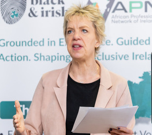 An individual speaks passionately while holding papers, standing in front of a banner with logos and text, conveying a formal and engaging tone.