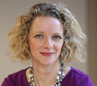 A woman with curly blonde hair, wearing a purple top and a beaded necklace, smiles softly against a neutral background, conveying warmth and professionalism.